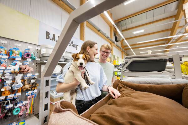 Eine Frau hält einen Hund im Tierfachmarkt, während sie zusammen mit einem Mann ein braunes Hundebett betrachtet.