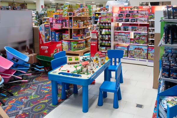 Du siehst einen Spielwarenbereich mit blauem Kindertisch, Spielzeughäusern, bunten Regalen und Schubkarren.