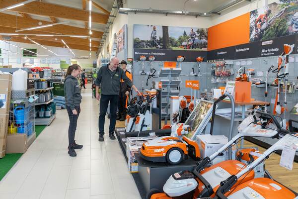 Du siehst zwei Personen im Raiffeisen-Markt vor einer Verkaufsfläche mit STIHL Gartengeräten und Rasenmähern.
