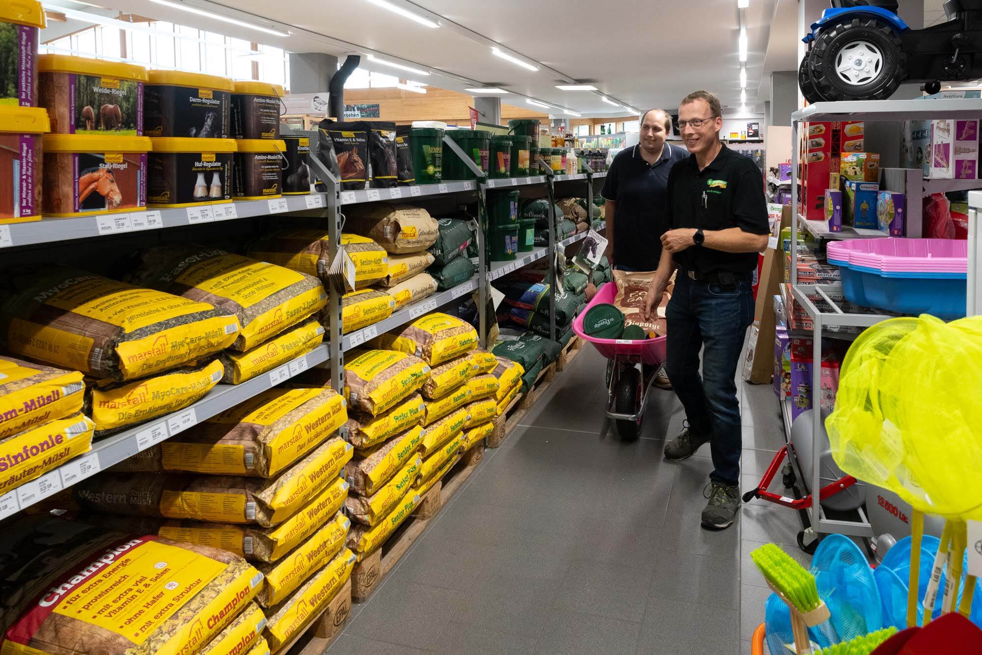 Zwei Männer schieben im Tierbedarfsladen eine pinke Schubkarre mit Futtersäcken an Regalen mit Pferdefutter vorbei.