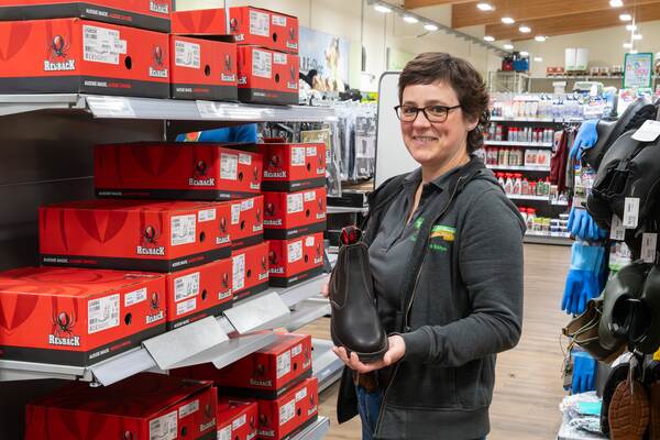 Du siehst eine Frau im Raiffeisen-Markt, die einen schwarzen Redback-Arbeitsschuh vor roten Kartons hält.