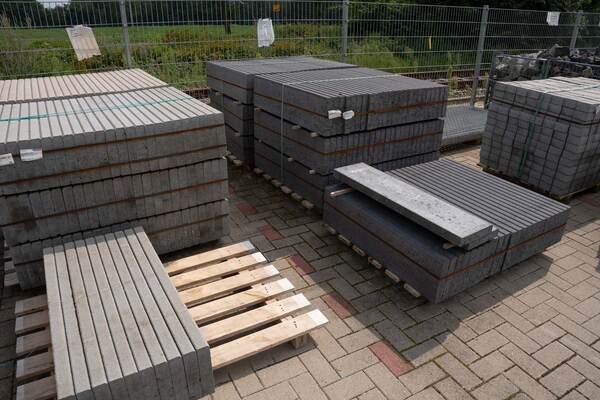 Du siehst gestapelte Paletten mit grauen Betonborden und Pflastersteinen auf einem Lagerplatz.