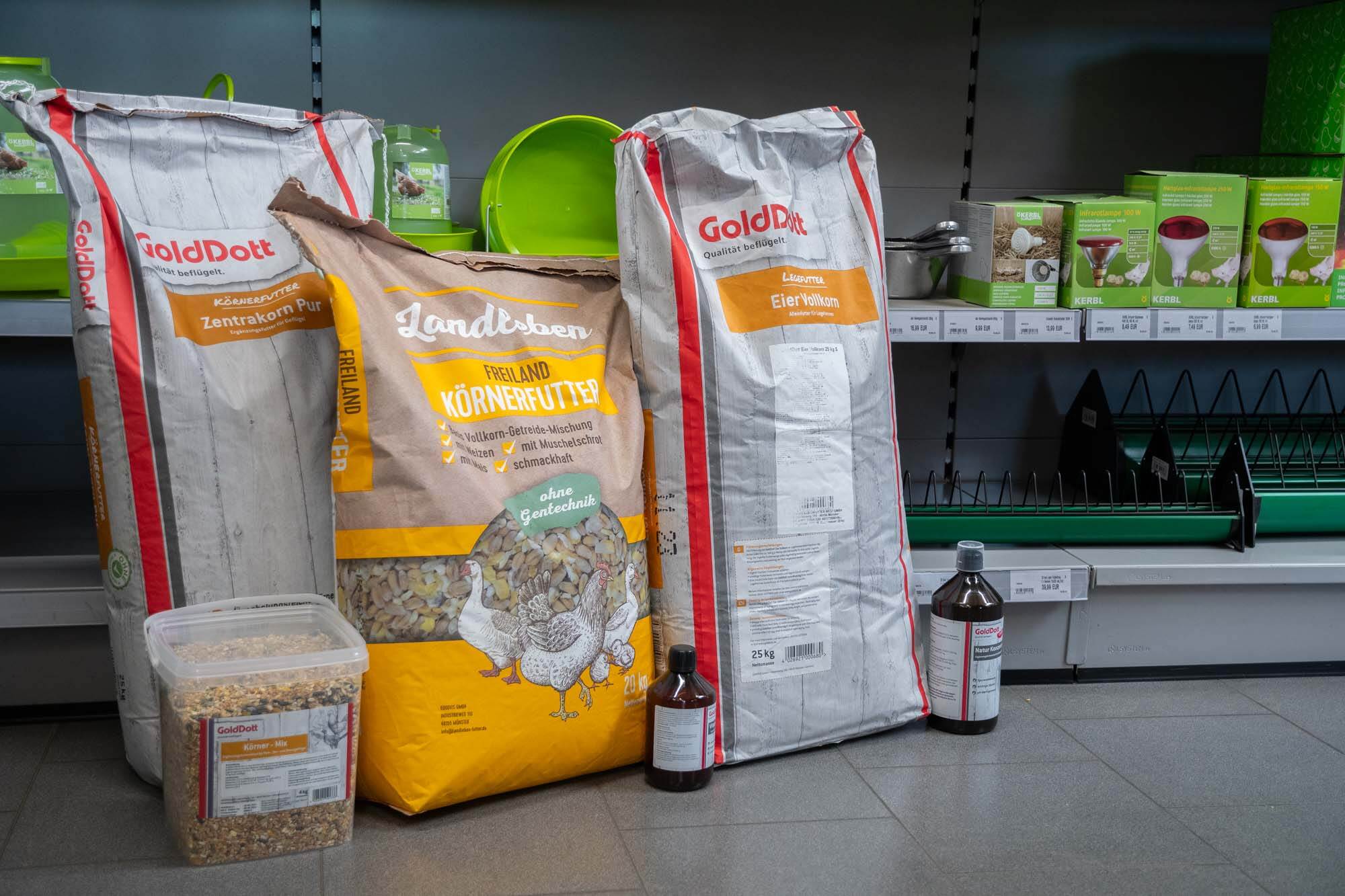 Große Säcke und ein Eimer mit Geflügelfutter stehen neben zwei Flaschen vor einem Regal im Tierbedarfsladen.