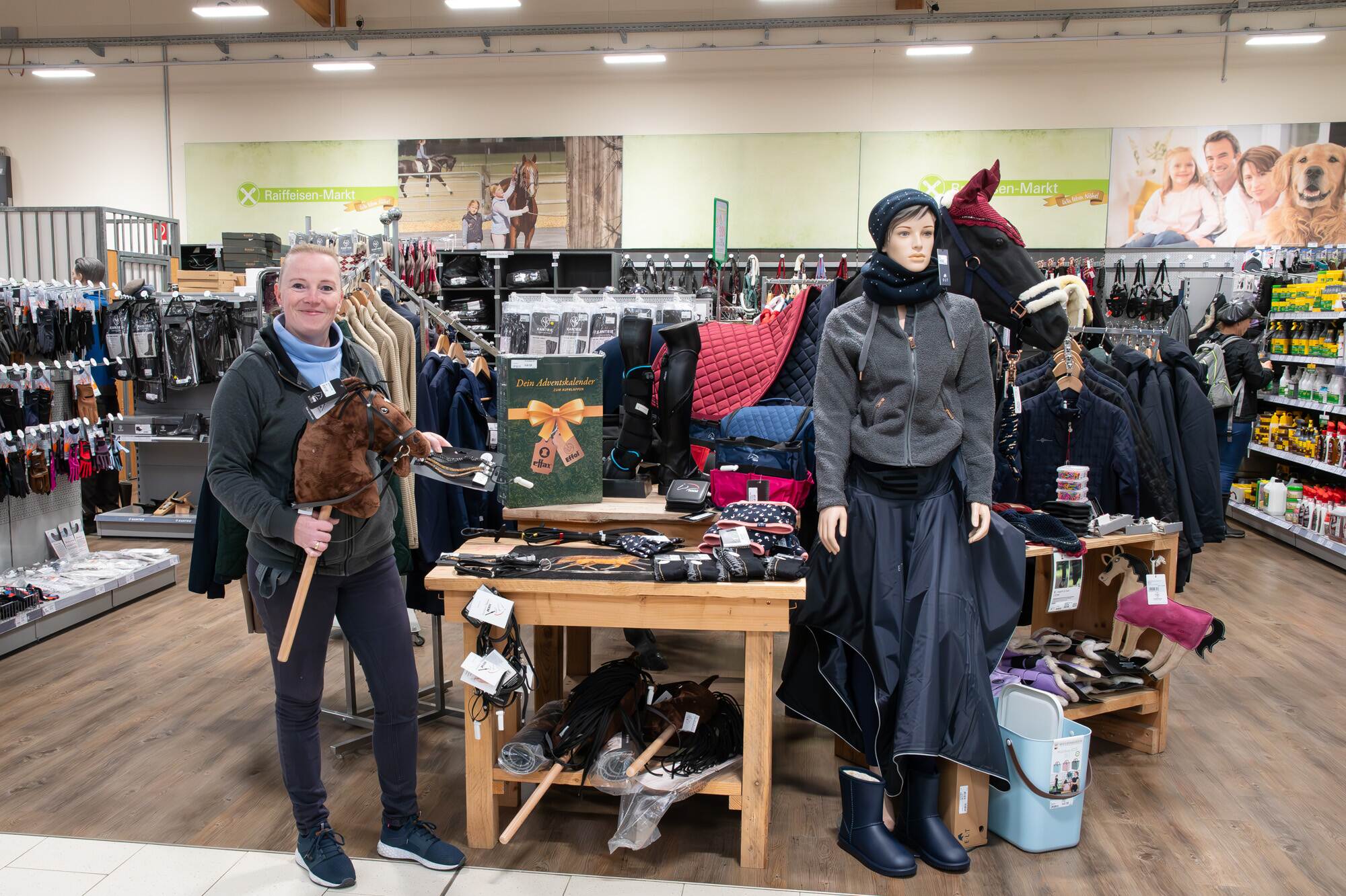 Frau in Reitsportabteilung hält ein Steckenpferd, neben einem Tisch mit Handschuhen und einer Schaufensterpuppe.