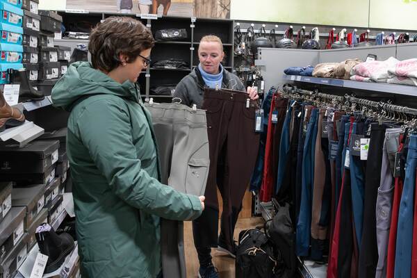 Zwei Frauen in einem Reitsportgeschäft vergleichen Reithosen vor einer Kleiderstange mit weiterer Reitbekleidung.