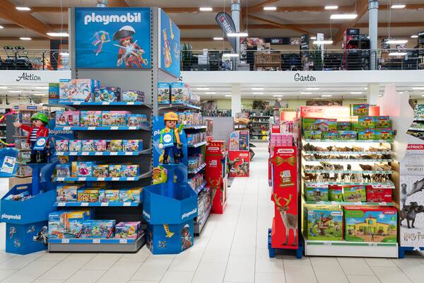 Du siehst ein Spielwarenregal mit Playmobil-Sets links und Schleich-Tierfiguren rechts in einem Geschäftsgang.