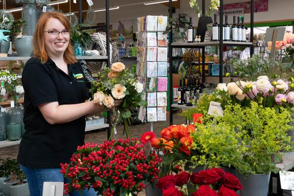 Du siehst eine Floristin im Raiffeisen-Markt, die einen Blumenstrauß zwischen frischen Schnittblumen bindet.