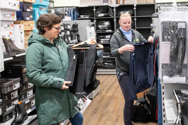 Zwei Frauen in einem Reitsportgeschäft betrachten Schutzwesten und Kleidung neben Regalen mit Stiefeln.