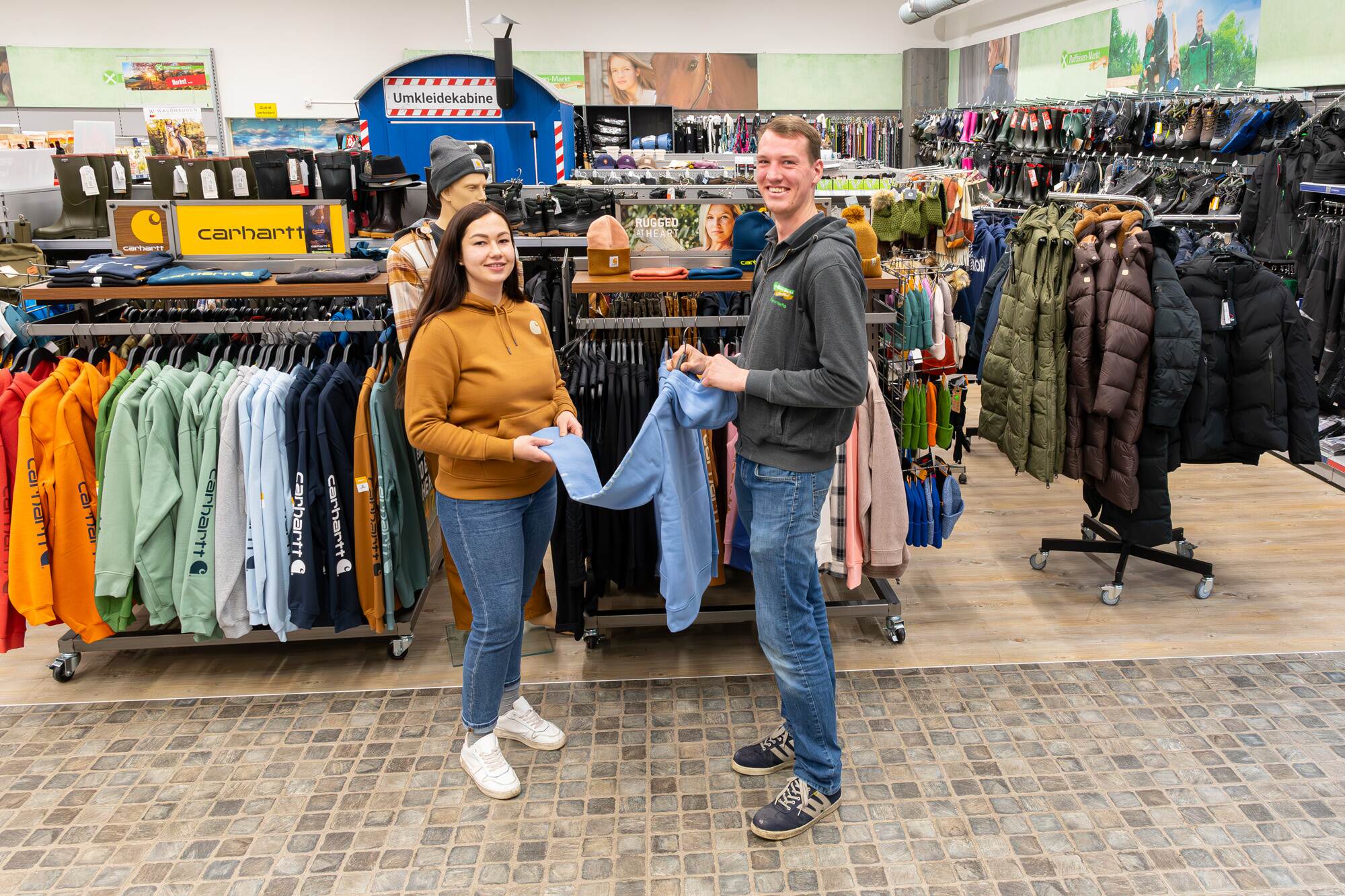Du siehst zwei Personen, die im Raiffeisen-Markt einen blauen Hoodie bei einer Auswahl von Carhartt Kleidung ansehen.
