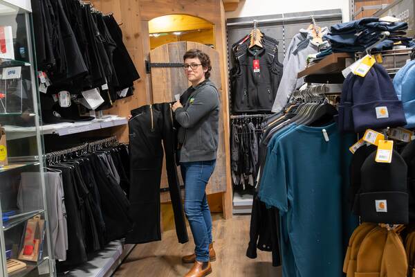 Du siehst eine Frau im Raiffeisen-Markt, die schwarze Arbeitshosen vor einer Umkleidekabine prüft.