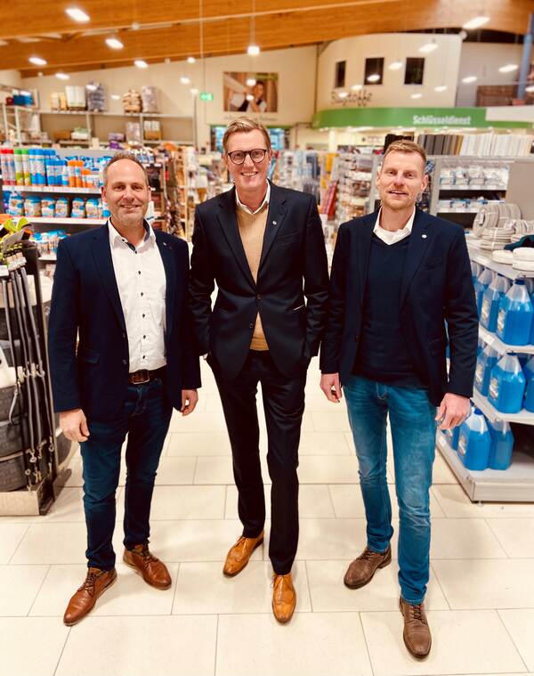 Du siehst drei Männer in Anzügen, die in einem Baumarkt zwischen Regalen für ein Foto posieren.