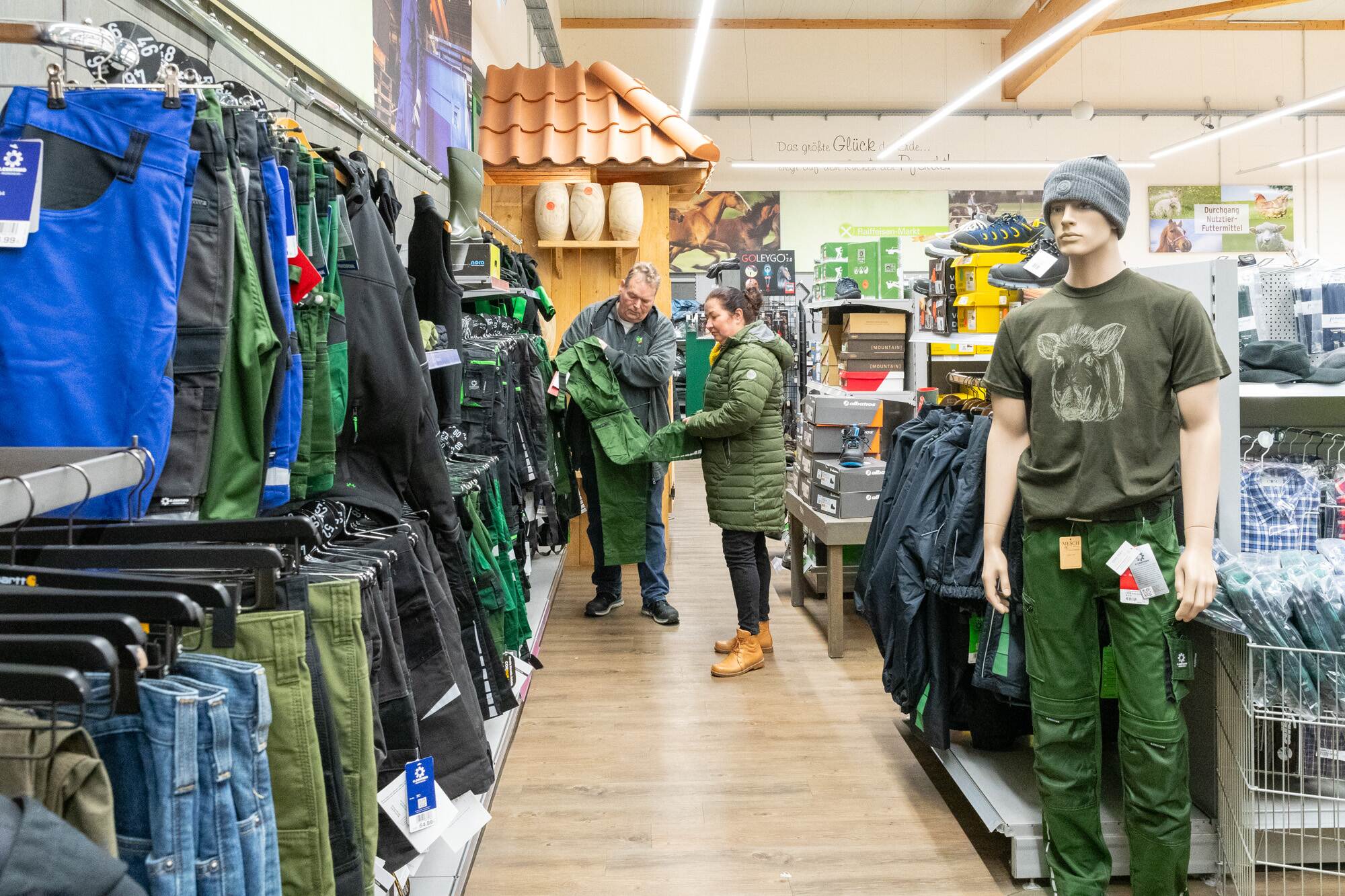 Du siehst zwei Personen im Raiffeisen-Markt, die grüne Arbeitskleidung ansehen, umgeben von Bekleidung und Schuhen.