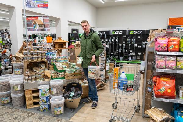 Ein Mann steht im Tierbedarfsgeschäft und hält einen Eimer mit Meisenknödeln vor einem Regal mit Vogelfutter.
