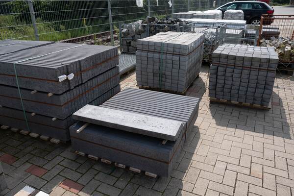 Du siehst gestapelte Paletten mit Bordsteinen, Pflastersteinen und Natursteinen auf einem Lagerplatz.