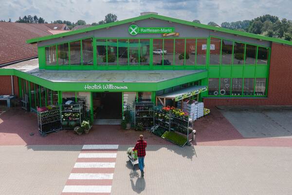 Du siehst den Haupteingang vom Raiffeisen Markt Gnarrenburg mit Pflanzen, Erden und einem Kunden mit Einkaufswagen.