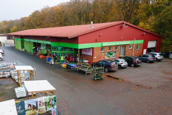 Außenansicht eines Raiffeisen-Marktes mit grünem Eingangsbereich, Schubkarren, Pflanzen und geparkten Autos davor.