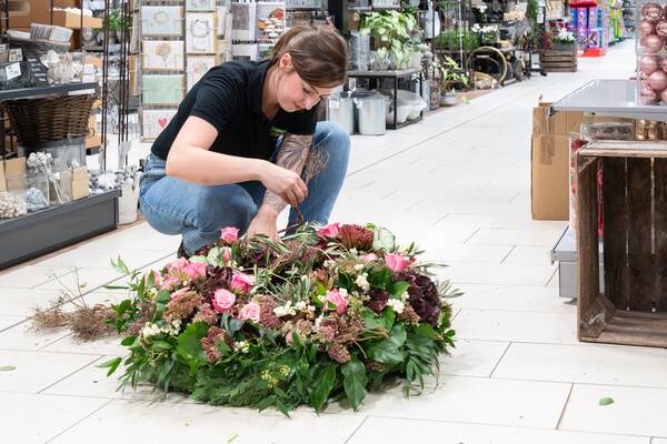 Du siehst eine Floristin im Raiffeisen-Markt, die am Boden einen großen, farbenfrohen Kranz bindet.