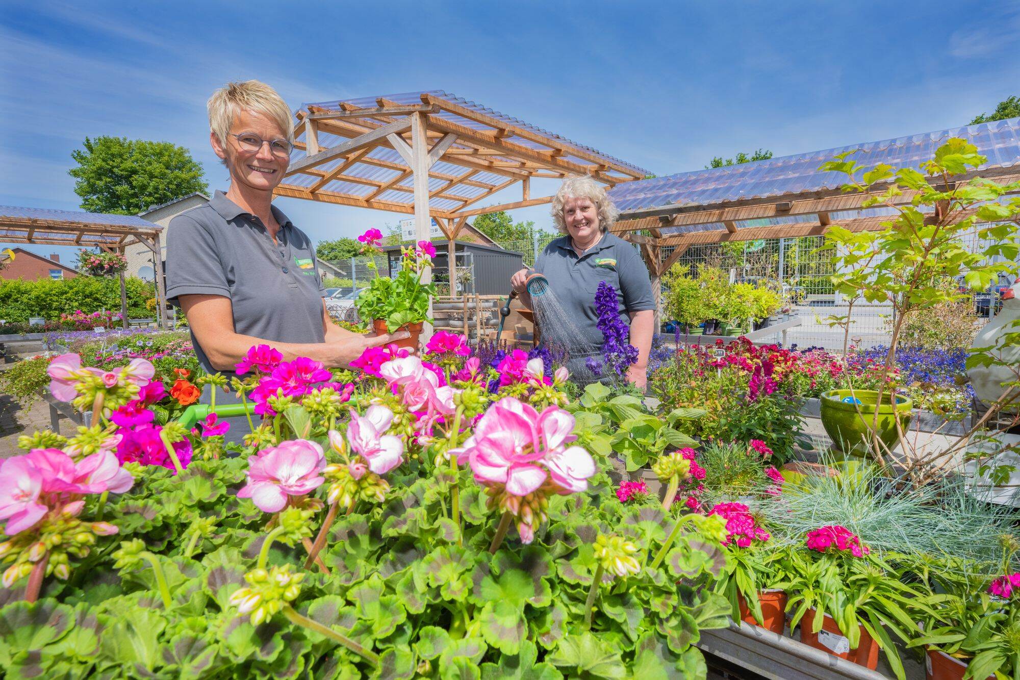 Zwei Floristinnen in einem Gartencenter umgeben von blühenden Sommerpflanzen, eine hält eine Gießkanne.