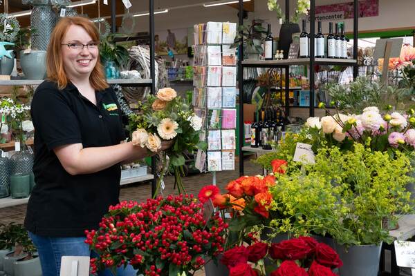 Du siehst eine Floristin im Raiffeisen-Markt, die einen Blumenstrauß zwischen frischen Schnittblumen bindet.