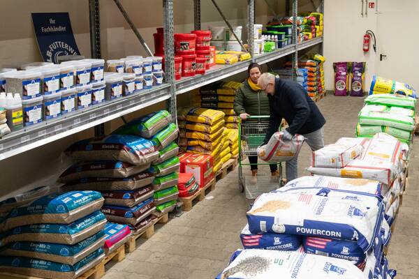 Mann lädt großen Sack Tierfutter in einen Einkaufswagen, während Frau daneben in einem Lagergang wartet.