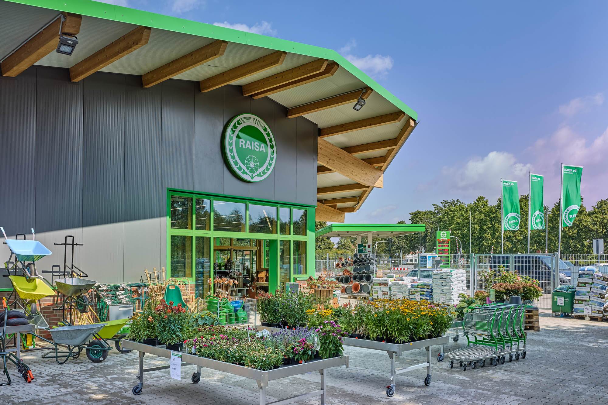 Du siehst den Eingangsbereich eines RAISA-Markts mit Blumen, Gartenbedarf und Schubkarren im Außenbereich.