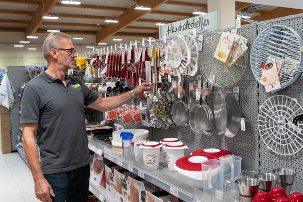 Du siehst einen Mann im Raiffeisen-Markt vor einem Regal mit Küchen- und Haushaltshelfern.