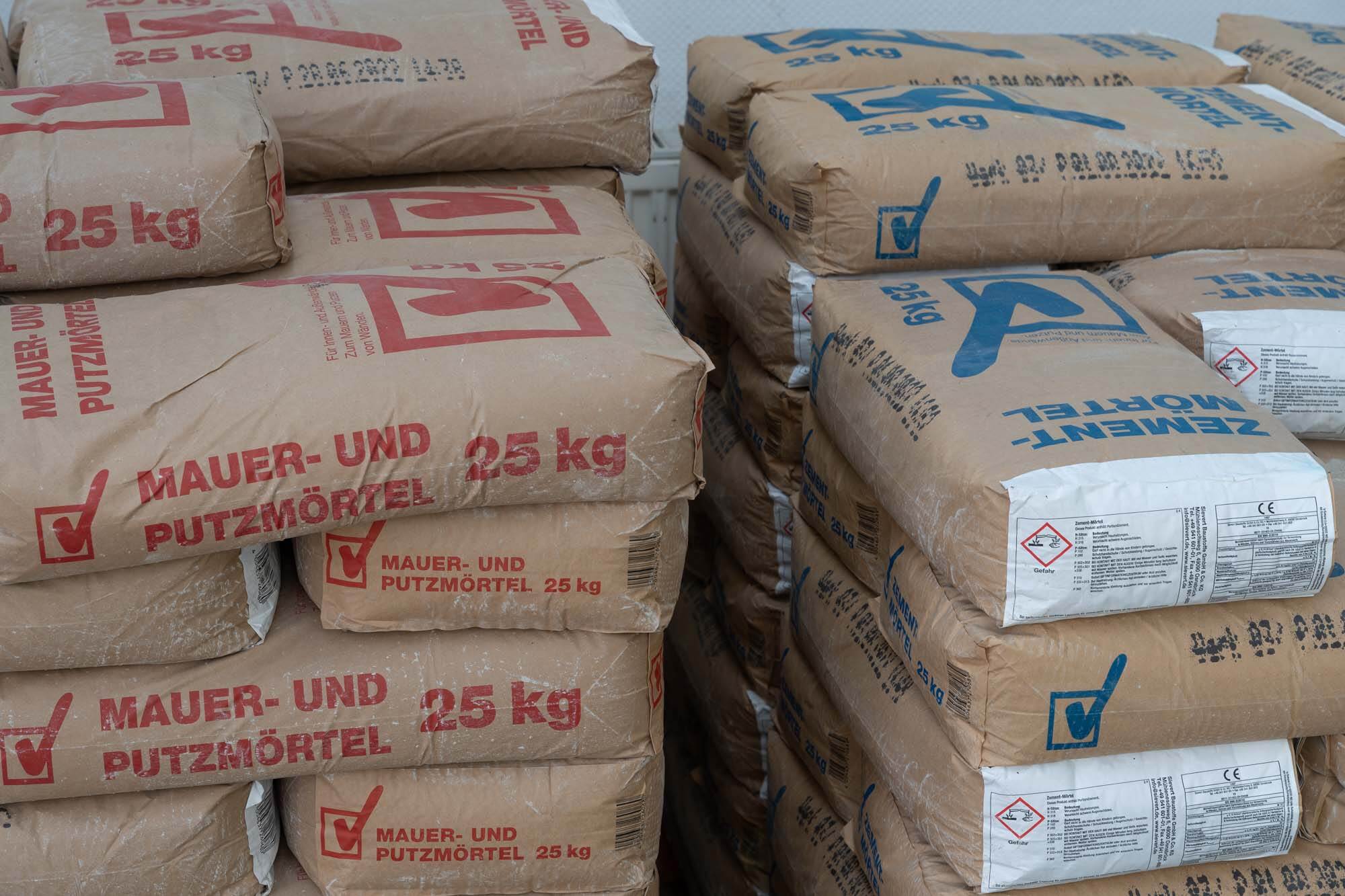 Du siehst gestapelte Säcke mit Mörtel und Zement in 25-kg-Verpackungen in einem Baumarkt.