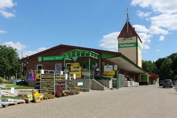 Eingang eines Raiffeisen-Marktes mit grünem Vordach, Pflanzen, Gartenartikeln und einem Turm im Hintergrund.