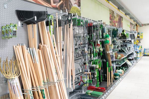 Du siehst ein Regal im Raiffeisen-Markt mit Gartenwerkzeugen wie Rechen, Hacken und Holzstielen.